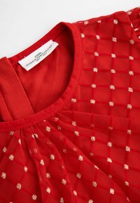 Czerwona sukienka z nakładką z przezroczystej tkaniny w kratę, zdobiona metalicznymi złotymi akcentami. Posiada okrągły dekolt i jednolitą czerwoną podszewkę.