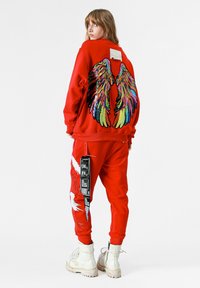 Sudadera roja con alas de lentejuelas en arcoíris en la parte posterior, combinada con joggers rojos que presentan gráficos de aves blancos y una etiqueta negra. Botas blancas.