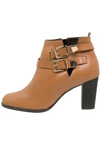 Bottine en cuir tan avec un talon empilé et deux sangles dorées à boucle à l'avant, ainsi que des découpes sur le côté.