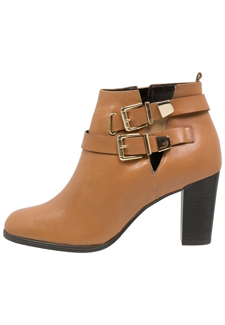 Bottine en cuir tan avec un talon empilé et deux sangles dorées à boucle à l'avant, ainsi que des découpes sur le côté.