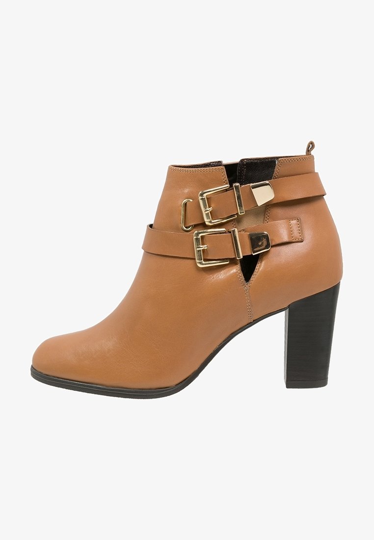 Bottine en cuir tan avec un talon empilé et deux sangles dorées à boucle à l'avant, ainsi que des découpes sur le côté.