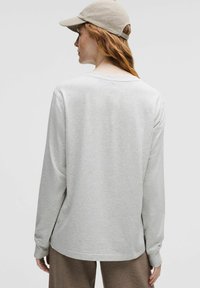 Lys grå langærmet skjorte med afslappet pasform, rund halsudskæring og blød tekstur. Kombineret med en beige kasket, set fra ryggen.