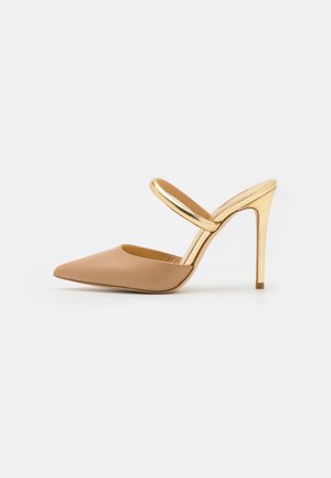 Beige mule med spids tå og høje hæle, dekoreret med en skinnende guldrem og stilethæl mod en hvid baggrund.
