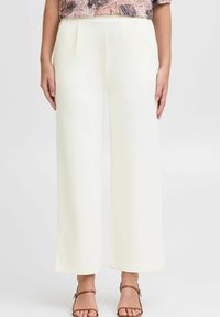 Pantalons évasés en tissu crème clair, avec des plis à la taille et une texture lisse. Associés à des sandales marron.