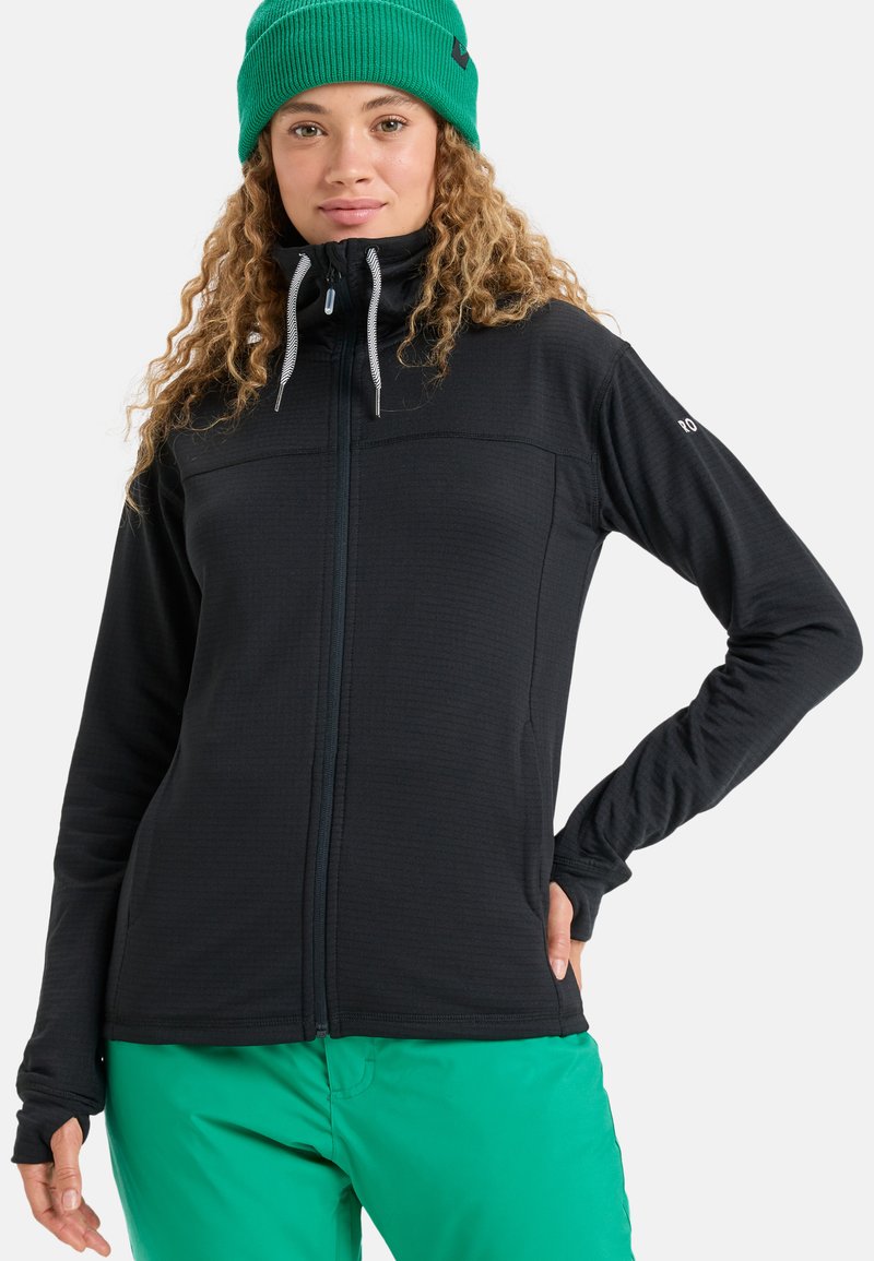 Čierna fleece bunda s plným zipsom vpredu, dvoma náprsními vreckami a reflexnými šnúrkami. V páre so zelenými nohavicami a zelenou pletenou čiapkou.