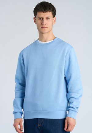Junger Mann steht frontal, trägt einen hellblauen langärmligen Pullover über einem weißen Unterhemd und dunkelblaue Jeans.
