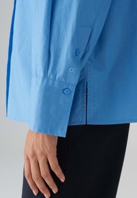 Camicia button-up azzurro chiaro con maniche lunghe, dotata di due bottoni blu ai polsini e di un piccolo logo sulla manica.