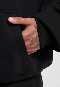 Svart hettegenser med teksturert stoff, frontlomme og ribbestrikkede mansjetter. En tatovert hånd er plassert i lommen, og viser detaljer.