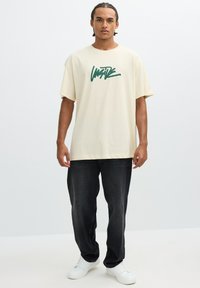 Camiseta de algodón color crema con un diseño gráfico verde, de manga corta y corte holgado. Llevada con jeans oscuros y zapatillas blancas.