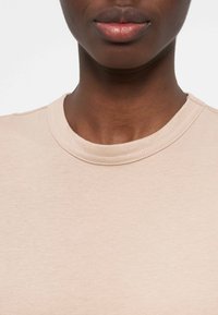 Κοντινό πλάνο ενός ατόμου με σκουρόχρωμο δέρμα που φοράει μπεζ μπλούζα με στρογγυλή λαιμόκοψη, που δείχνει τον λαιμό και τα χείλη του πάνω σε απλό φόντο.