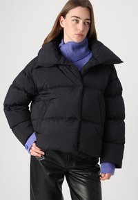 Sort pufferjakke med bred krave og store quiltede sektioner. Bæres over en lys lilla turtleneck, parret med sorte læderbukser.