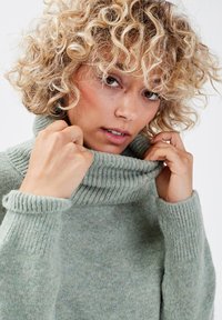 Suéter verde de cuello alto hecho de un tejido suave y texturizado. Puños y cuello de canalé. El modelo ajusta el cuello con ambas manos.