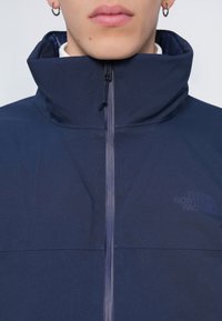 Homme portant une veste North Face bleu marine zippée avec un col montant et des boucles d'oreilles argentées en forme d'anneau, visible sur fond blanc.