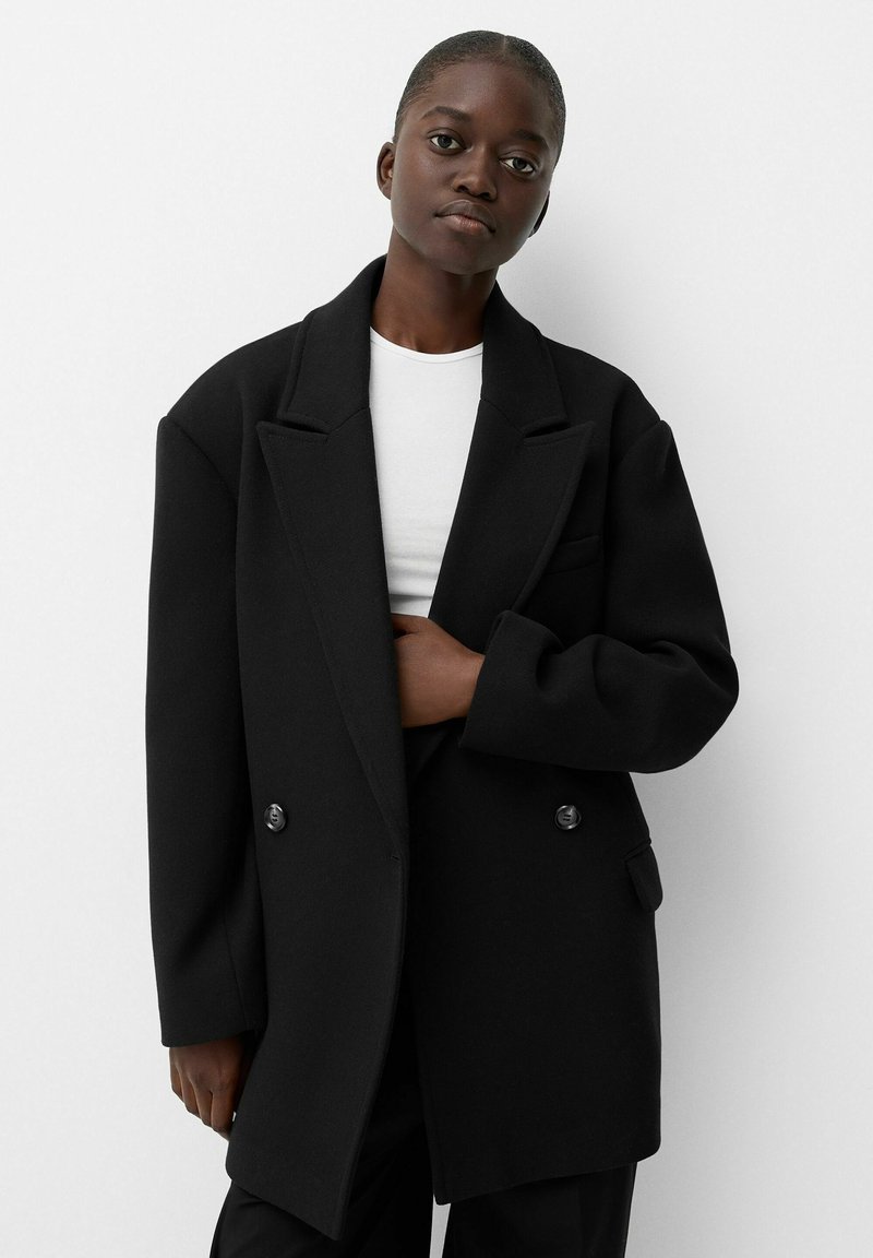 Schwarzer, doppelt knöpfbarer Mantel aus dickem Stoff, mit übergroßen Revers, großen Knöpfen und einem lässigen Schnitt. Darunter ein schlichtes weißes T-Shirt.