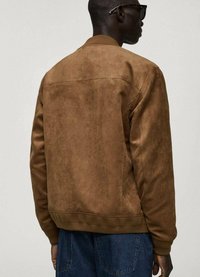 Giacca bomber in suede marrone con finitura testurizzata. Presenta un colletto e polsini a costine, cucitura del pannello posteriore e vestibilità rilassata. Indossata con jeans blu.