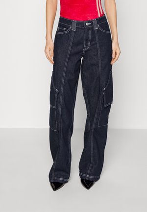 Femme portant un jean cargo bleu foncé à jambes larges avec des coutures blanches, associé à des chaussures noires pointues et un haut rouge sans manches.