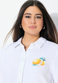 Camicia bianca in lino con una finitura testurizzata, caratterizzata da un motivo di limone ricamato colorato sul lato sinistro del petto. Chiusura con bottoni e colletto.