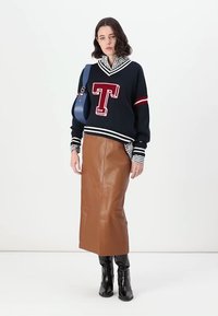 Maglione a maglia blu con accenti bianchi e rossi, caratterizzato da un grande logo "T", abbinato a una gonna midi in pelle marrone e stivaletti neri.
