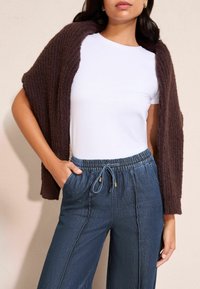 Barna kötött stóla fehér póló felett, sötétkék, széles szárú nadrággal párosítva, amely rugalmas derékpánttal és húzózsinórral rendelkezik.