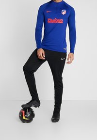 Sininen pitkähihainen urheilupaita, jossa on neljännesvetoketju, ja siinä on punainen sponsorilogo sekä seuran emblem. Mustat urheiluhousut, joissa on valkoinen logo ja yksityiskohtia.