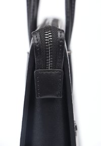 Sorte lædertaske, der viser en tekstureret side, med en metallisk lynlås og flad remtilslutning. Vertikal orientering med minimal detaljer.