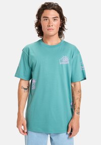 Tyrkysové bavlněné tričko s krátkými rukávy, zdobené růžovými grafickými motivy, včetně slunce a palmy, a uvolněným střihem.