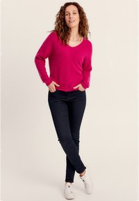 Un pull rose vif avec un design en V, associé à un jean bleu foncé et des baskets blanches. Le pull possède une texture douce et duveteuse.