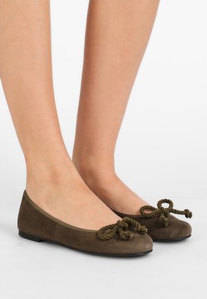 Brune ruskind ballet flats med detaljer af snoet rebsløjfe, båret på bare fødder mod en ensartet hvid baggrund.