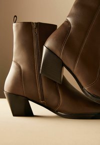 Bottines en cuir brun avec fermeture éclair sur le côté, talon carré et détails de couture. Texture lisse, bout rond et design minimaliste.
