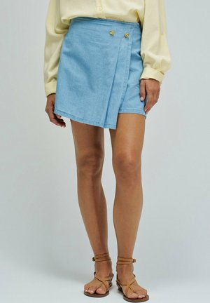 Mujer que lleva una falda cruzada azul claro con dos botones dorados, blusa beige y sandalias marrones con tiras, de pie frente a un fondo liso.