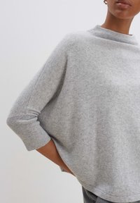 Světle šedý, měkký pletený svetr s vysokým límcem, spadlými rameny a rukávy délky 3/4, s volným střihem a minimálním po�čtem švů.