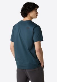 Teal kortærmet t-shirt lavet af blødt stof, med rund halsudskæring og et sort logo på øvre ryg. Parret med mørke bukser.