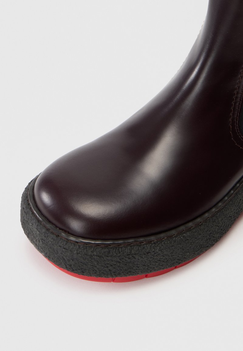 Stivaletto in pelle bordeaux con punta tonda, suola in gomma nera testurizzata e dettagli rossi visibili sul bordo della suola.