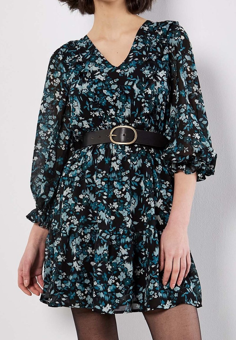 Femme portant une robe noire à fleurs bleues et blanches, manches bouffantes, et une ceinture noire avec une boucle ovale dorée.