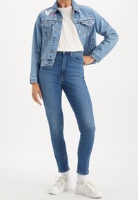 Chaqueta de mezclilla en azul claro con un bolsillo frontal, con detalles bordados de corazones. Jeans ajustados azules combinados con zapatillas blancas.