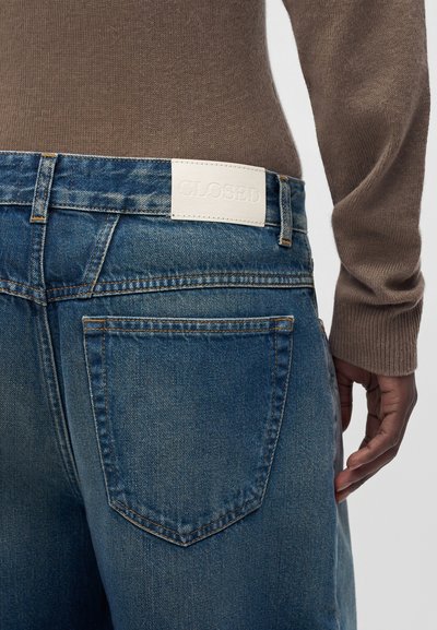 Gros plan de la vue arrière montrant un jean en denim bleu avec une étiquette blanche « CLOSED » et une personne portant un haut marron à manches longues.