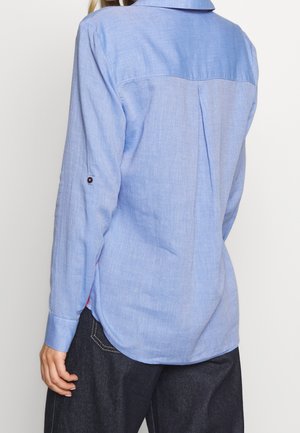 Personne portant une chemise à manches longues bleu clair avec des poignets boutonnés et un pantalon en denim foncé, vue de dos sur un fond uni.