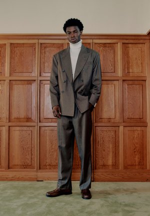 Homme portant un costume gris croisé, un col roulé blanc et des chaussures marron, debout contre un mur en panneaux de bois et un sol légèrement moquetté.