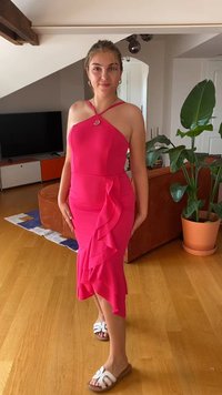Fuksijos rožinė suknelė su prigludusia siluetu, volanginių akcentų, kaklo juostos ir iki kelių ilgio. Dėvima su baltais sandalais.