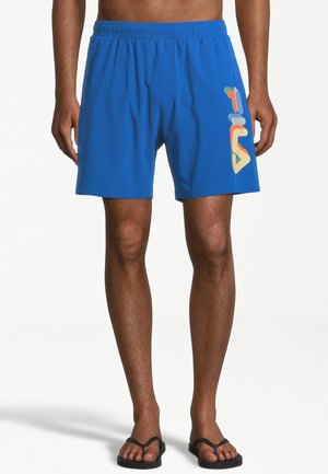 Homme portant un short bleu FILA avec un logo multicolore sur la cuisse droite, debout pieds en tongs noires sur fond blanc.
