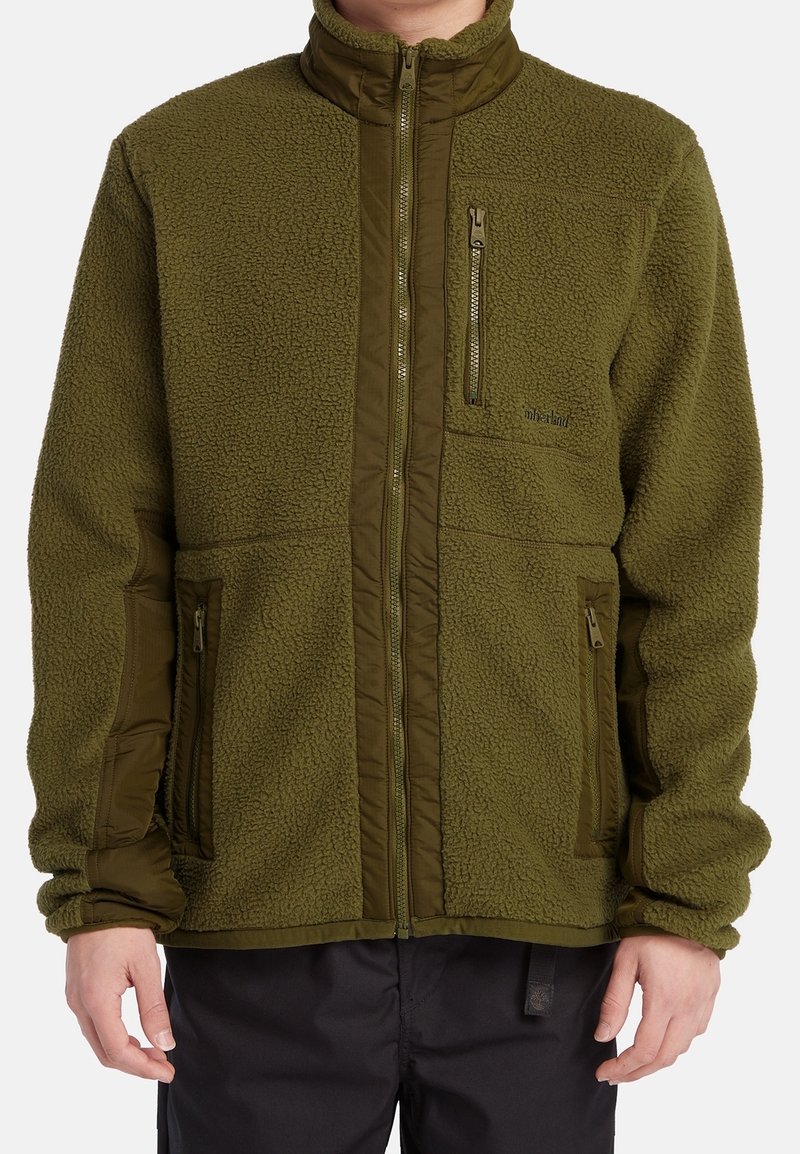 Homme portant une veste polaire vert olive avec des poches zippées devant et sur la poitrine, debout les mains détendues sur les côtés.