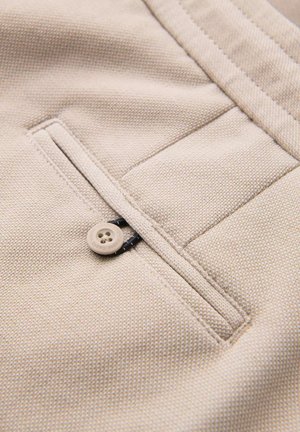 Beige fabric pocket featuring a button closure, seam detailing, and textured weave. The pocket is neatly stitched with contrasting thread.