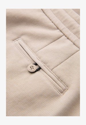 Beżowa kieszeń z tkaniny z zapięciem na guzik, detalami szwów i teksturowanym splotem. Kieszeń jest starannie wykończona kontrastową nitką.