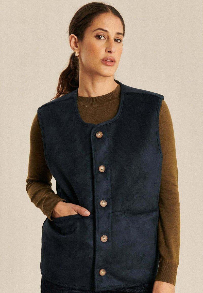 Marineblauwe, zachttextuur vest met een ronde hals, voorkant met knoopsluiting en twee zijzakken. Bruine knopen en een relaxte pasvorm.