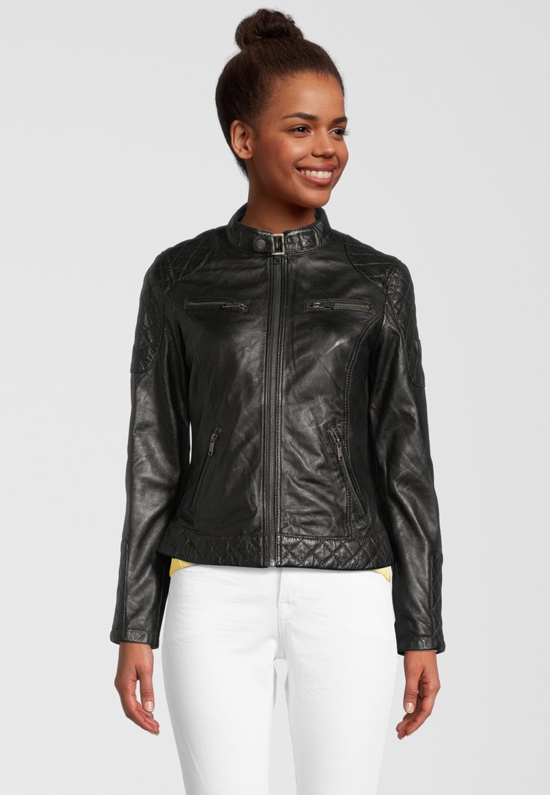 Zwarte leren jas met staande kraag, gestikte schouderaccenten, ritszakken en een slim-fit ontwerp. Witte broek zichtbaar eronder.