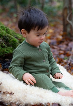Grüner Waffelstrick-Overall mit rundem Halsausschnitt, drei Holzknopfverschluss und langen Ärmeln, getragen von einem sitzenden Kind auf einer weichen, weißen Oberfläche.
