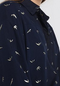 Camicia blu navy con colletto a punta, caratterizzata da piccoli motivi a foglia dorata sparsi, indossata da una persona con lunghi capelli castani.