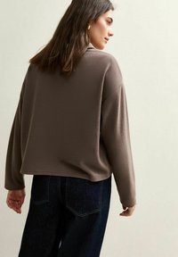 Brązowy sweter z teksturą, o luźnym kroju, z opadającymi ramionami i skróconym dołem, zestawiony z ciemnoniebieskimi dżinsami.