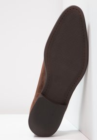 Chaussure en daim marron avec une semelle en caoutchouc noir lisse, présentant un fond texturé et des coutures contrastantes le long du bord.