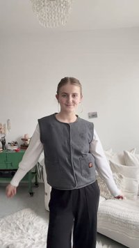 Graue ärmellose Weste mit zwei Vordertaschen und dekorativen Nähten, getragen über einem weißen Langarmhemd, kombiniert mit schwarzen Hosen.